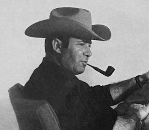 A portrait of Arthur Umanoff wearing a cattleman hat and smoking a tobacco pipe